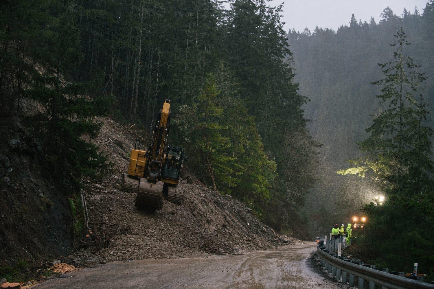 In a forested area, heavy construction equipment sits on a slope near a road containing debris.