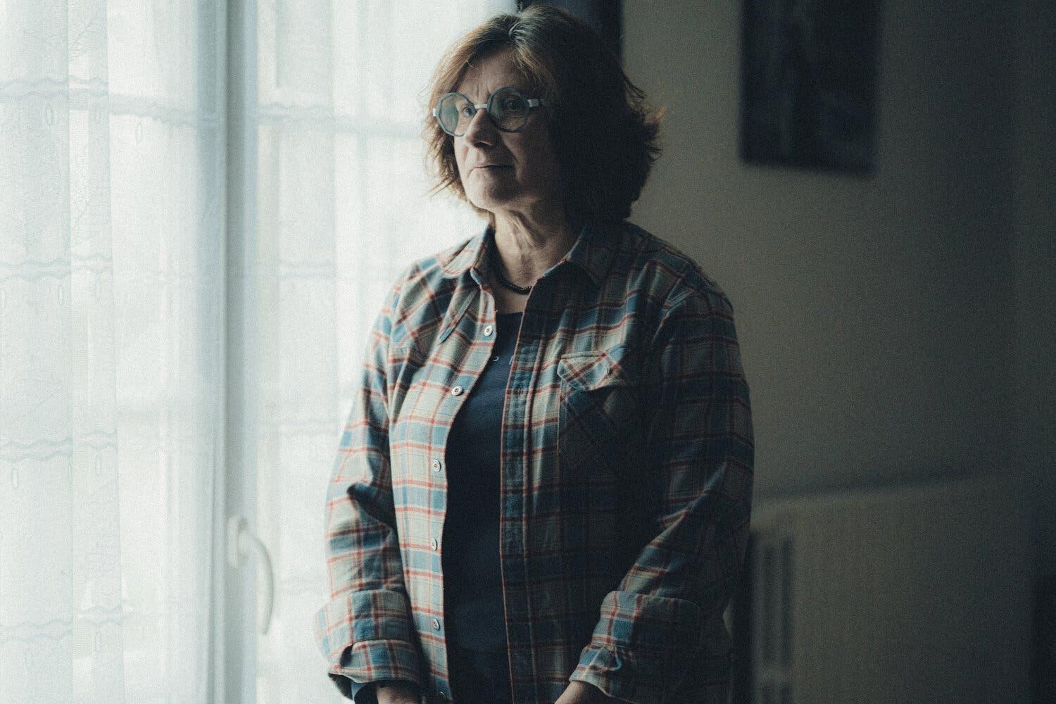 Christine Jagueneau, wearing a plaid shirt and glasses, stands in front of a window in her home in Liniez, France.