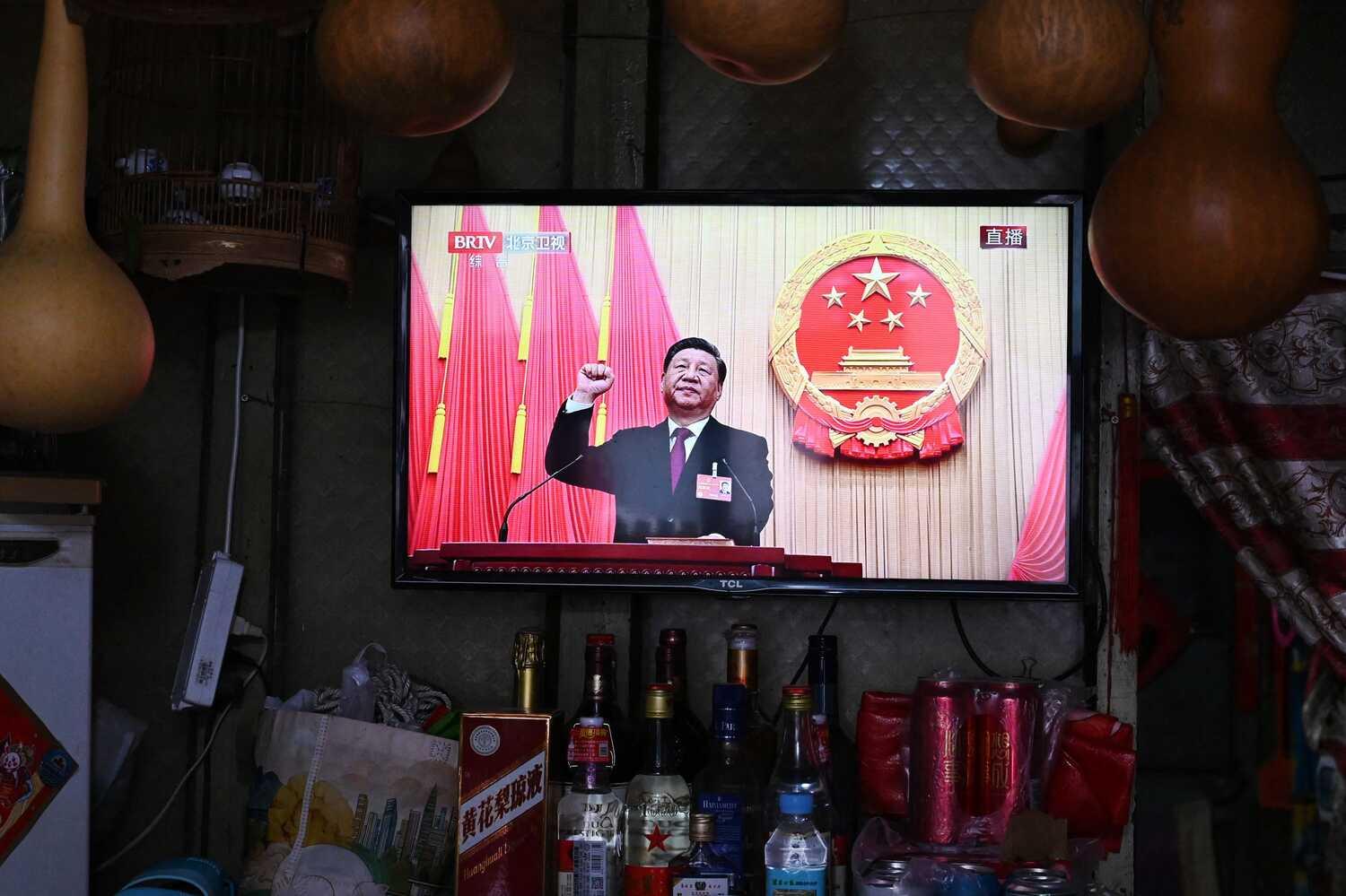 On a television screen, Xi Jinping, in a dark suit, with pink and beige curtains behind him. The bright TV screen contrasts with the dark background of goods for sale at a convenience store.