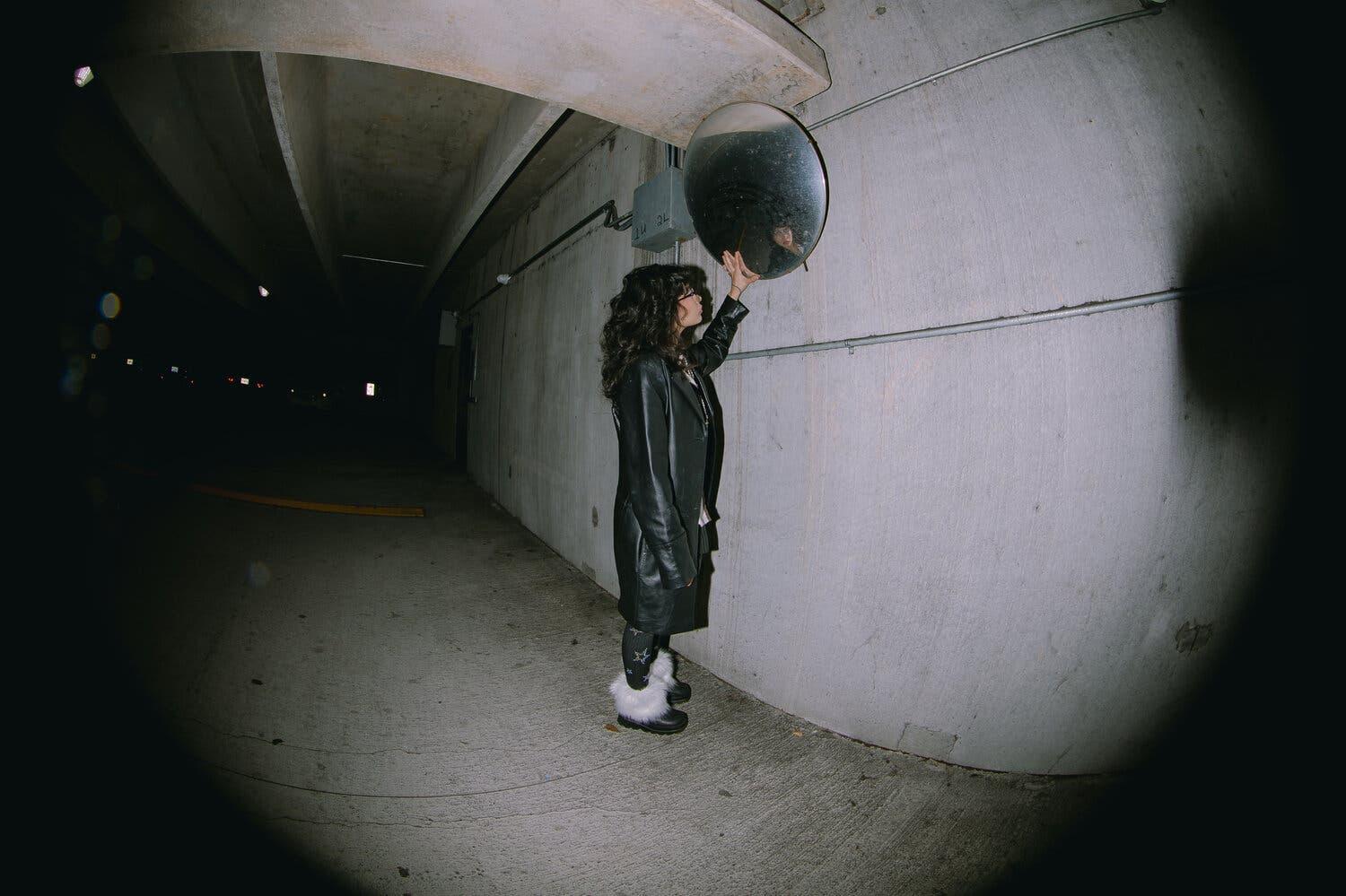 Mercedes Jimenez-Cortez, dressed in a long black leather coat, touches a concave traffic mirror in a dark parking garage.