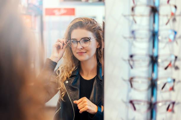 Smiling young woman trying on glasses on mirror in optician. Smiling young woman trying on glasses on mirror in optician. optical shop design stock pictures, royalty-free photos & images