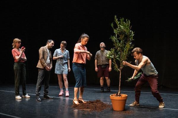 In a scene from the show “Are we not drawn onward to new era,” a man rips branches from a potted sapling. To his right are five actors, each doing something different: one woman is eating an apple; a man seems to be leaning into another woman; a woman is stepping in a mound of dirt; and a man is looking upward.