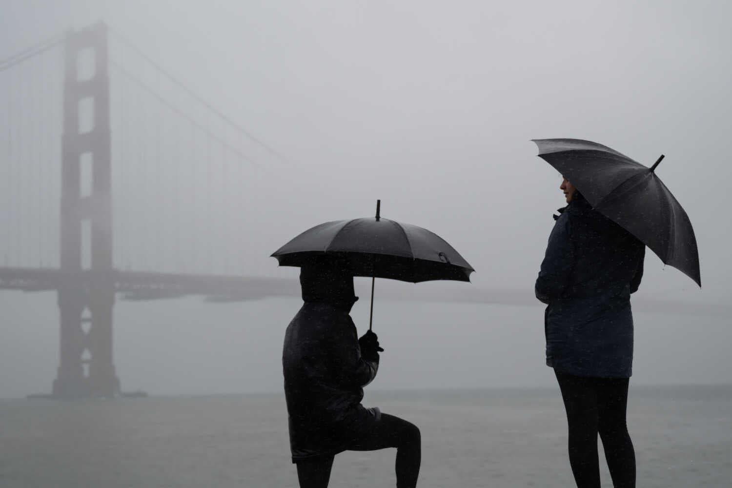 Two people, seen from behind and mostly in silhouettes, dressed in rain gear and holding umbrellas. In the distance, a large suspension bridge looms in the fog.