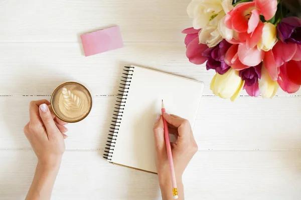 Overhead shot of woman hands drawing, writing with pencil in open notebook, drinking coffee on white wooden table. Beautiful tulip flowers bouquet of different colors. Background, copy space, close up Royalty Free Stock Photos