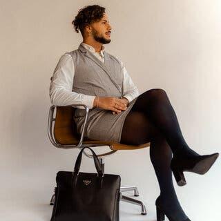 A person with curly brown hair and a dark beard sits in an office chair, legs crossed at the knee, in front of a white backdrop. He is wearing a gray sheath dress of sorts over a white button-up shirt; sheer black stockings; black pointy-toed heels; and several pieces of jewelry. A black Prada briefcase rests against the chair.