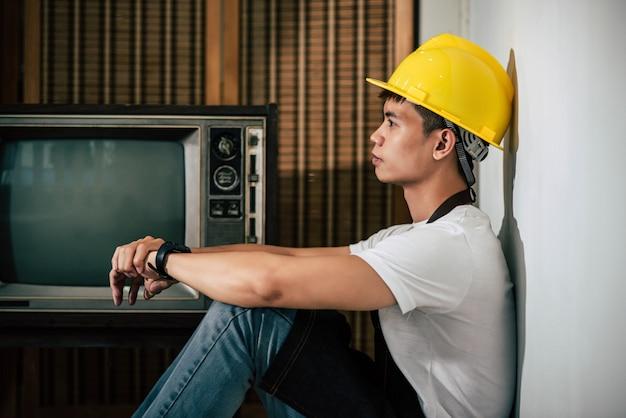 The mechanic is wearing a yellow hat and hands resting on the knees.
