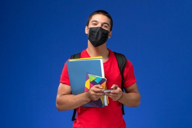 Front view male student in red t-shirt wearing backpack with mask holding files and using his phone on the blue background. Free Photo
