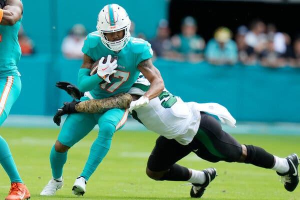 Wide receiver Jaylen Waddle, in the Dolphins’ teal uniform, carries the ball while being tackled by Jets linebacker C.J. Mosley, with his arms around Waddle’s waist.