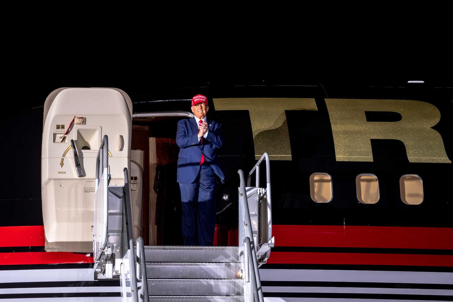 Donald Trump, wearing a red cap, stands at the exit door of his jet, which is painted in red, white and blue, with stairs in front of him. The letters T and R are visible behind him.