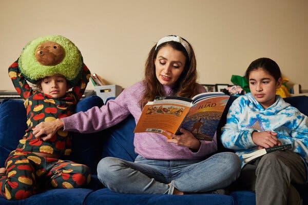 A woman, in a light purple top and jeans, is sitting on a blue couch and reading a book to two boys on either side of her. The one on her right is in colorful pajamas (with a stuffed avocado on his head) and the other one is in a white and blue sweatshirt. She appears to be tickling the boy to her right.