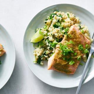 Two plates topped with a piece of salmon on a bed of rice are photographed from overhead. They’re scattered with scallion slices and plated alongside lime wedges.