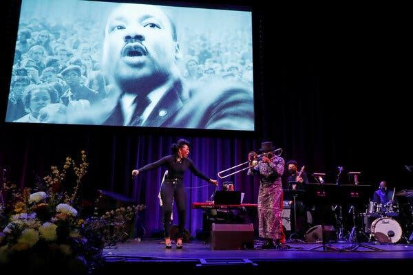 A woman dressed in black performs onstage with a band; above her is a screen with an image of the Rev. Dr. Martin Luther King Jr.