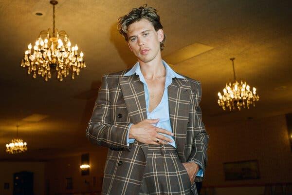 In a portrait, Austin Butler wears a brown plaid, wide-lapel suit over an open-collar blue shirt. He’s standing in a room with chandeliers and looking down at the camera with a neutral expression.