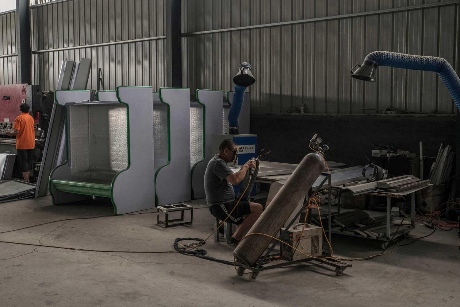 A factory floor that appears to be producing refrigerator cases for shops. At the center, a man with welding goggles sits at a bench adjusting a torch.