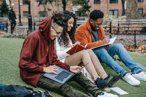 Happy multiracial group of students in casual clothes sitting together on grass and studying with laptop and books
