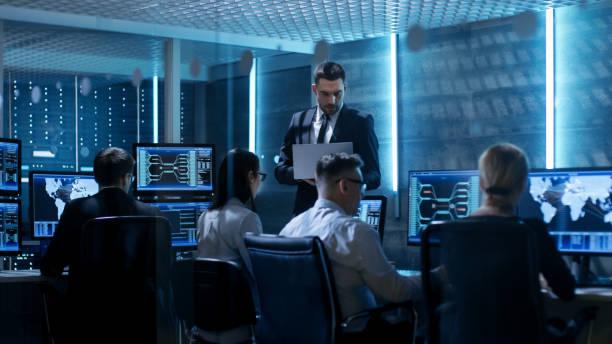 Professional IT Engineers Working in System Control Center Full of Monitors and Servers. Supervisor Holds Laptop and Holds a Briefing. Possibly Government Agency Conducts Investigation. Professional IT Engineers Working in System Control Center Full of Monitors and Servers. Supervisor Holds Laptop and Holds a Briefing. Possibly Government Agency Conducts Investigation. it support stock pictures, royalty-free photos & images