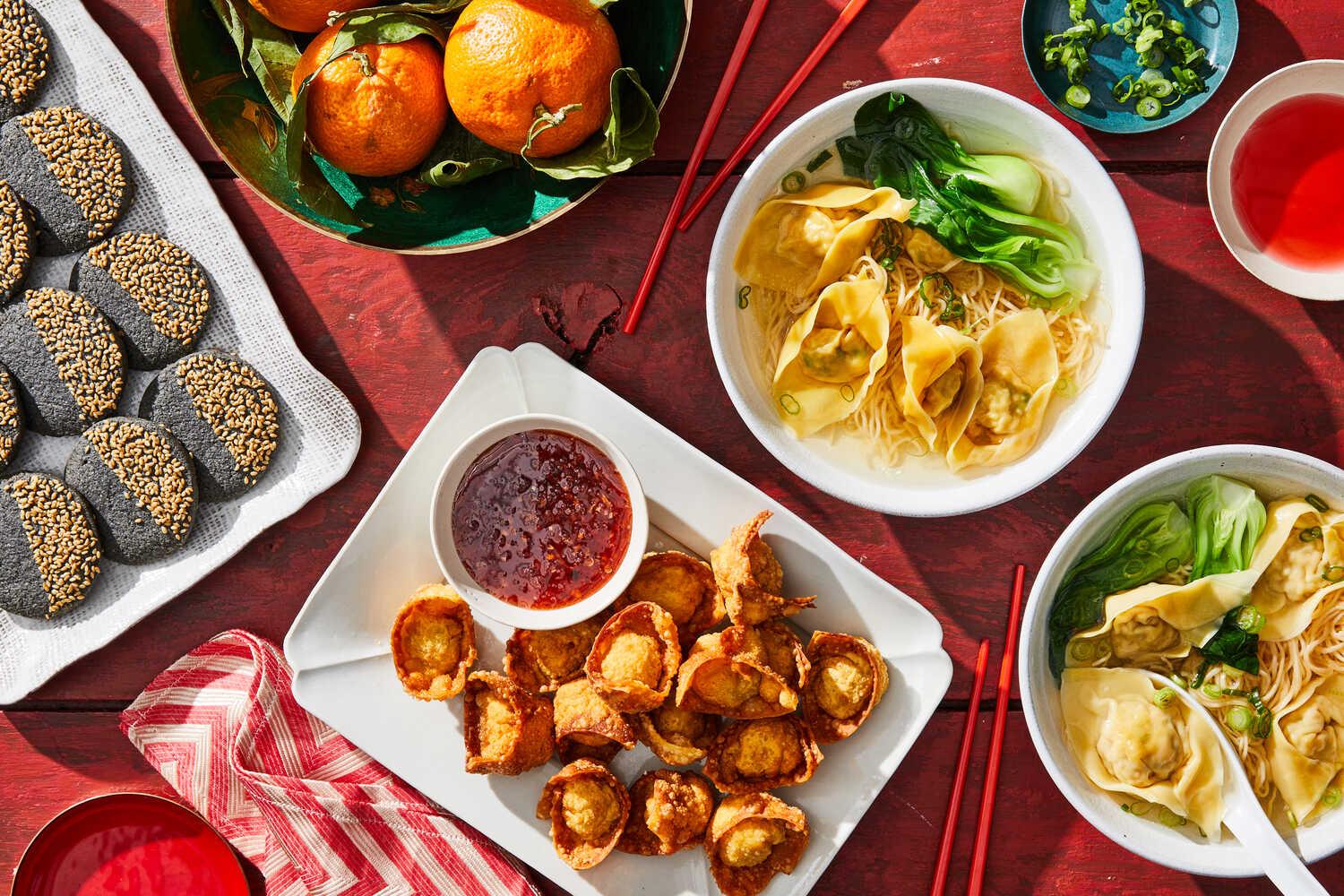 A Chinese New Year spread of food including black sesame cookies on a white platter, mandarin oranges in a bowl, fried wontons with dipping sauce on a plate and two bowls of wonton noodle soup.