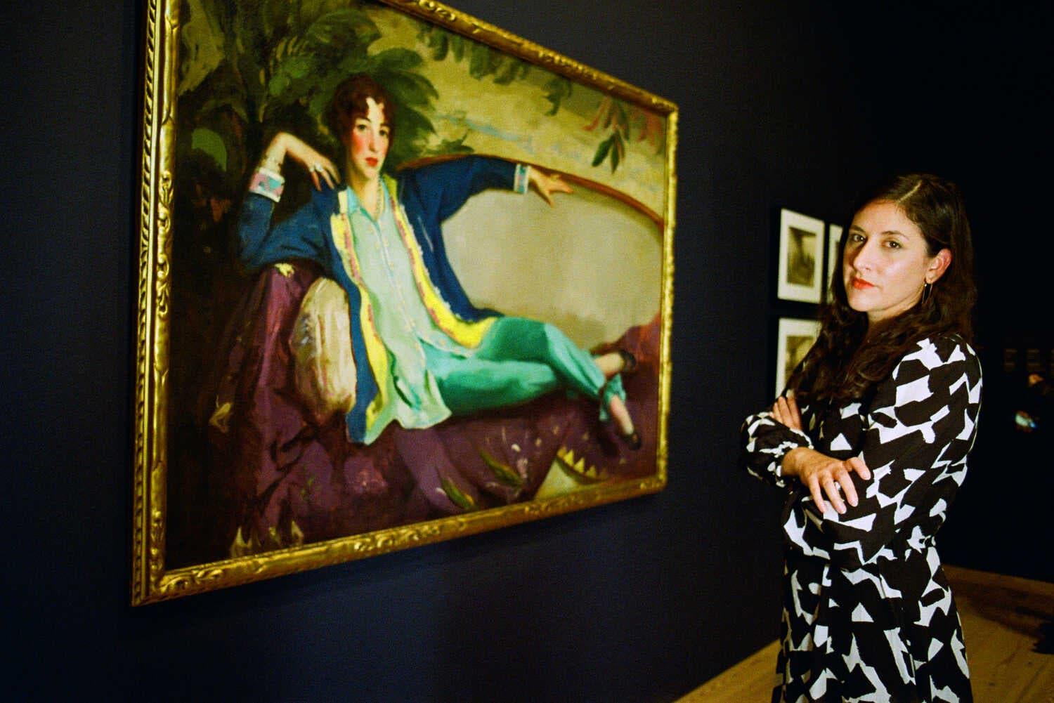 Marcela Guerrero, an associate curator at the Whitney Museum of American Art, stands in a print dress, with folded arms, next to a painting of Gertrude Vanderbilt Whitney.