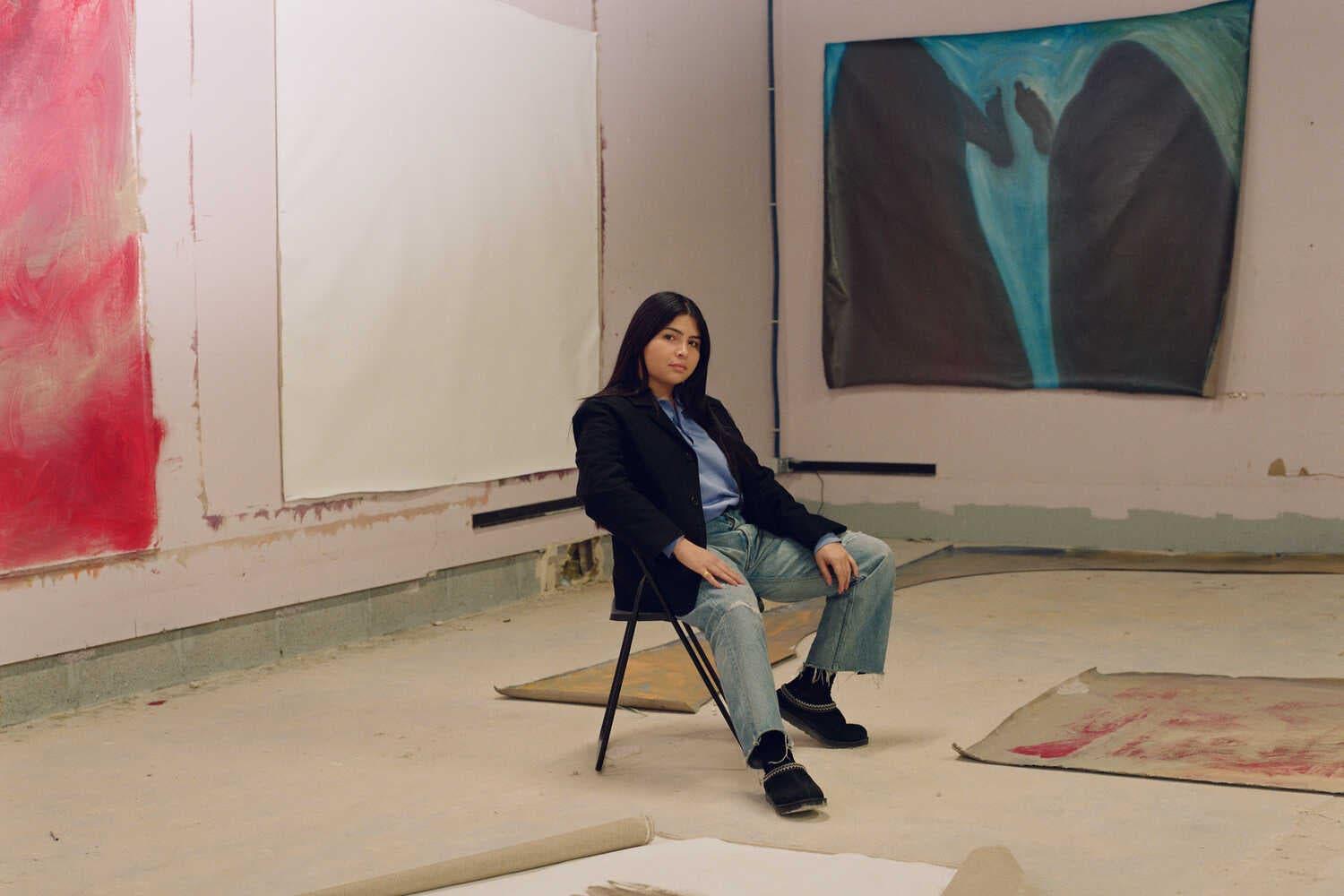The artist and poet Ser Serpas, in jeans and a dark jacket, sits before her expressive new paintings, in red and blue, from her ongoing series depicting faceless torsos and abstract bodies. Some are self-portraits.