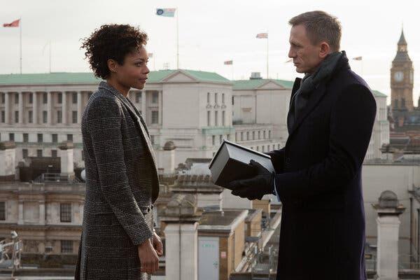A man in a black overcoat, gloves and gray scarf, and a woman in a tweed coat face each other as they stand on a rooftop. The man is holding a gray box. A large stately building with a green roof, columns and flags flying atop it dominates the background. To the right is an ornate clock tower, and below the people are more urban buildings.