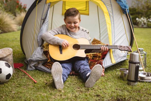 Caucasian boy smiling while playing guitar sitting in a tent in the garden Caucasian boy smiling while playing guitar sitting in a tent in the garden. childhood and hobby concept Party Rentals stock pictures, royalty-free photos & images