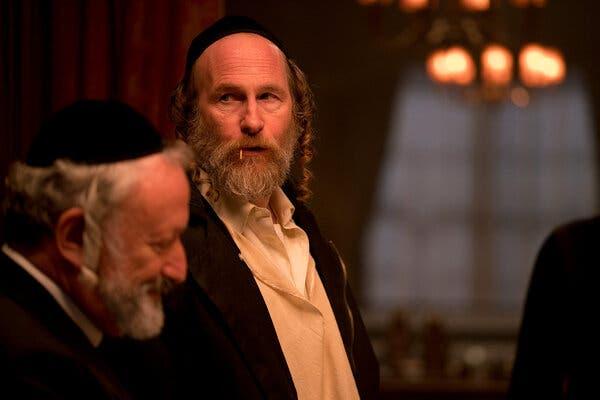 Two men in Hasidic garb stand at the left of the frame in a warmly lit room, with one out of focus in profile and the other looking off to the side with a toothpick in his mouth. A third man stands to the right, facing the camera and looking downcast.