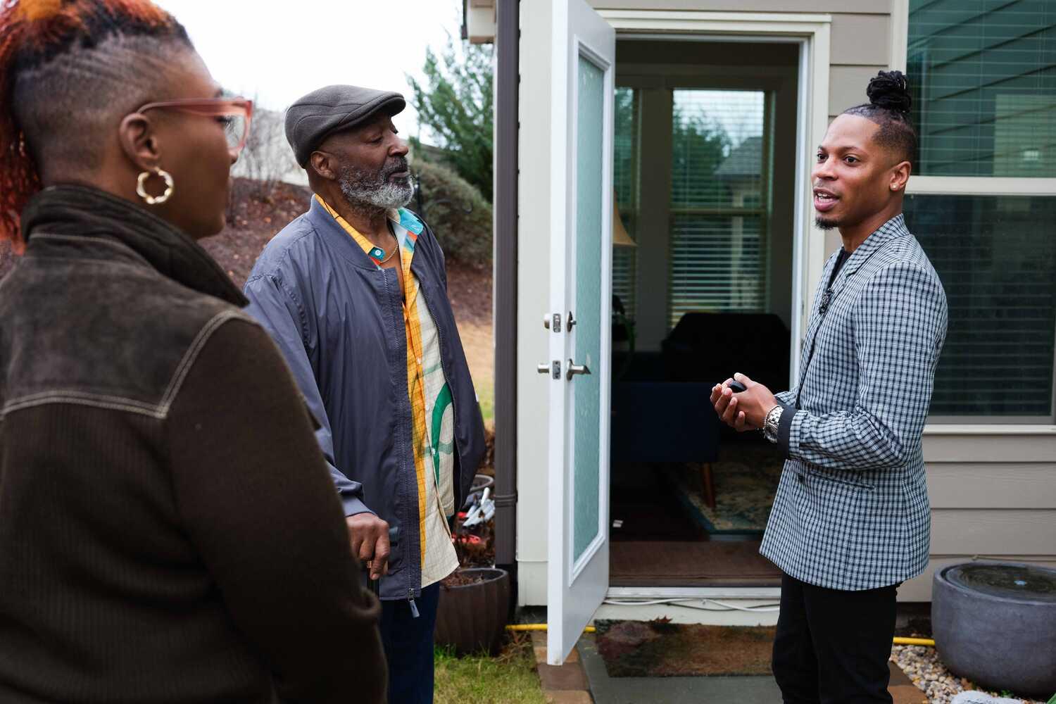 A man wearing a checkered jacket and dark-colored pants stands outside a house and talks to a man and woman. The other man is wearing a leather flat cap, a colorful button-down shirt and a blue jacket. He has a gray beard. The woman has a partially shaved head and reddish-orange locks.