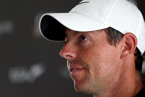 Rory McIlroy, shown in a white ball cap in a close-up shot, listens to a reporter’s question during a news conference.
