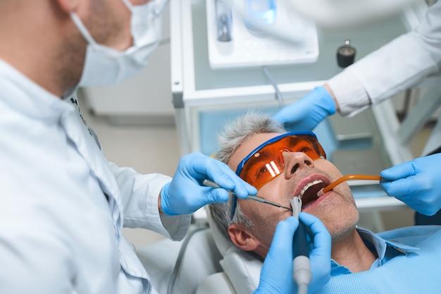 Male is lying in dental chair while dentist and assistant treating him root canal with modern equipment