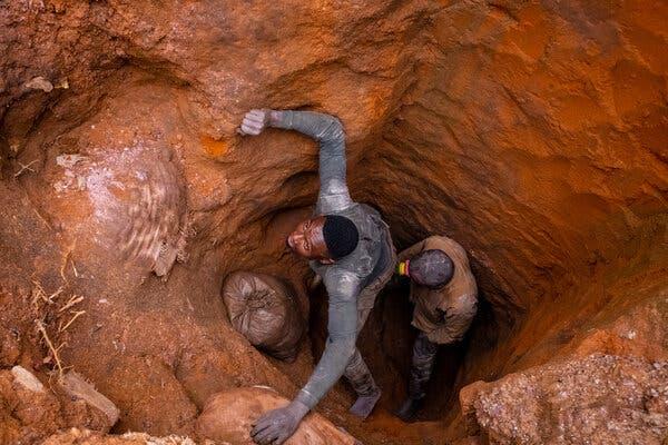 Miners look up from within a deep pit dug from reddish-brown earth.