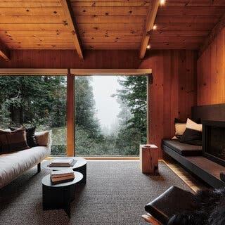 A living room paneled in pine, with a simple gray carpet, a light-colored sofa on the left, several simple, low stools in the center of the room and a minimalist, black wood stove on the right side. Straight ahead are two large floor-to-ceiling windows overlooking the forest.