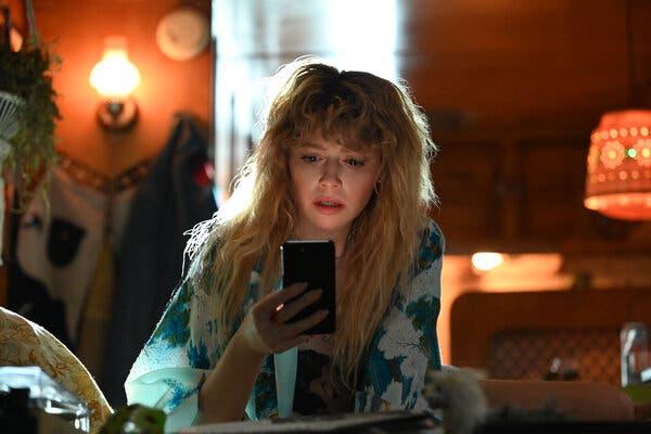 Natasha Lyonne staring at a phone screen in a scene from “Poker Face.” She is looking worried about or alarmed by what she sees on the screen. She’s sitting in a room that has orange overtones.