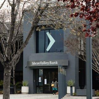 A gray building has a large logo in the upstairs window which looks like a light blue greater-than sign. Above the door, the name Silicon Valley Bank is written in white on a gray sign. Trees with red leaves are in the foreground of the photo.