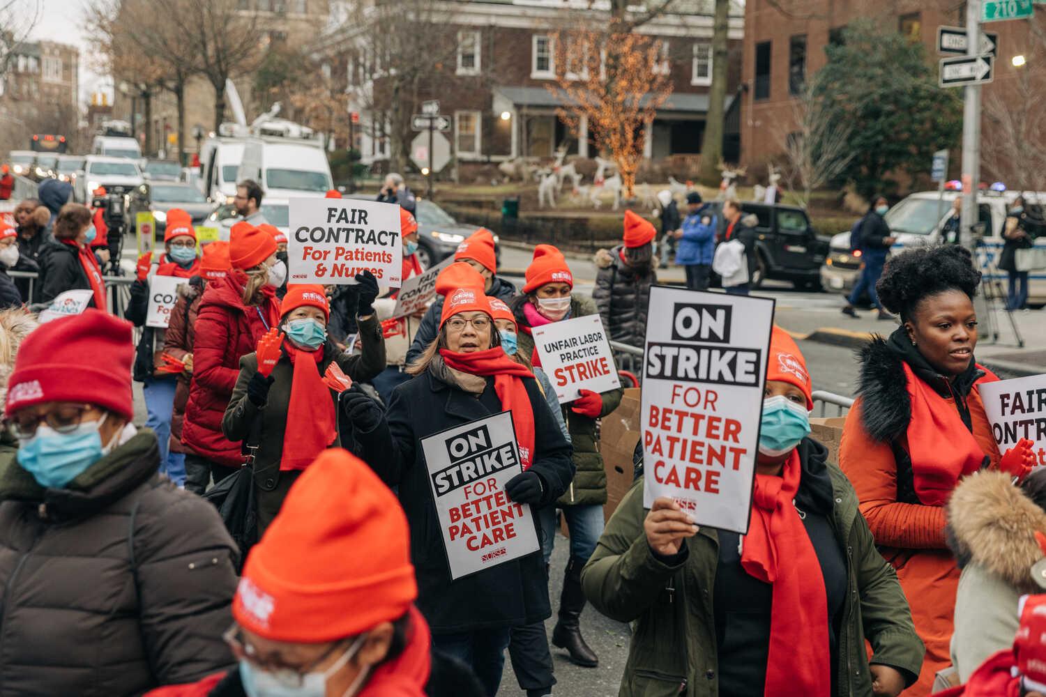 Women wearing red hats, scarves and gloves march down a sidewalk holding signs that say, “On strike for better patient care.”