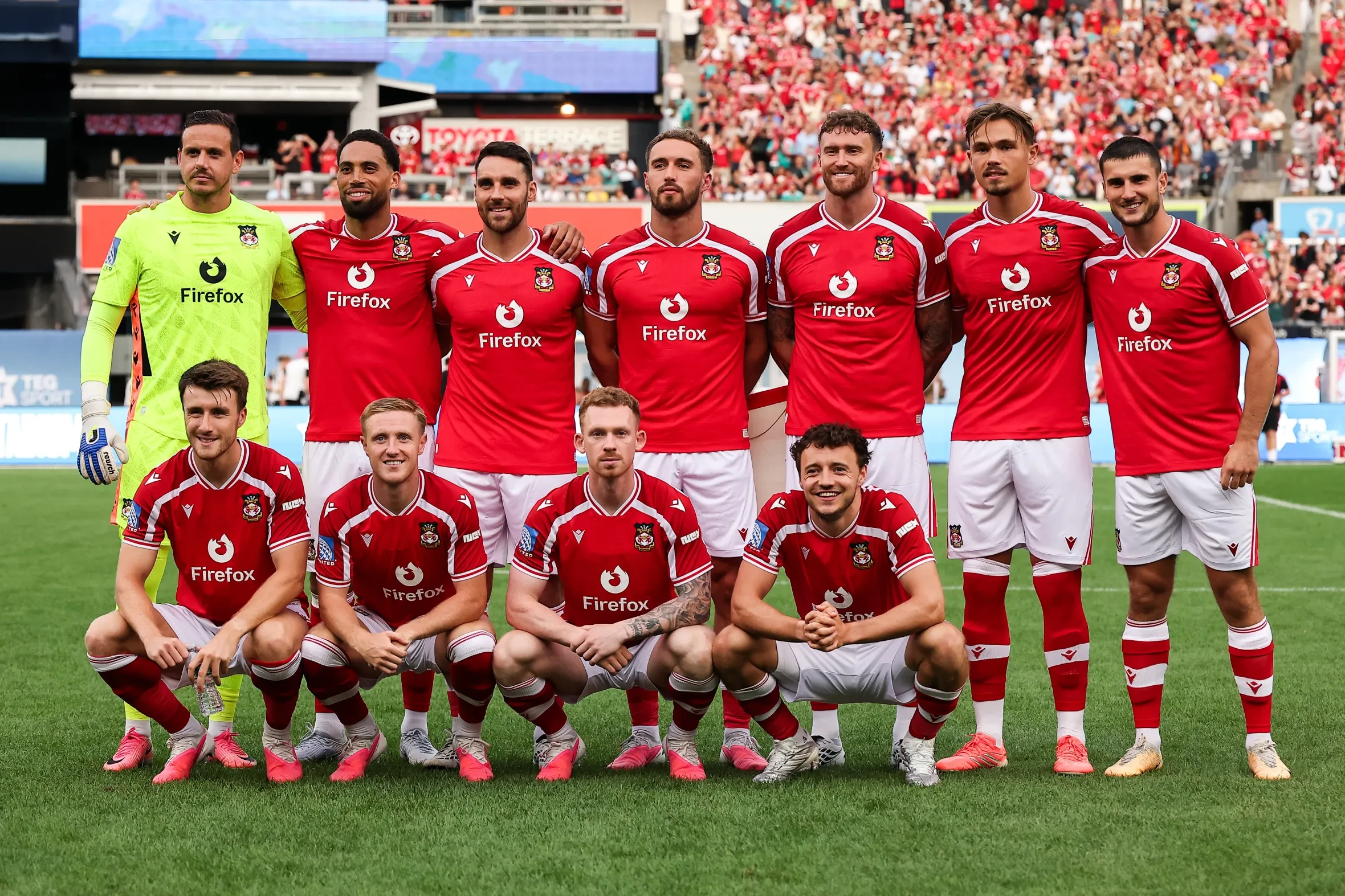 The Wrexham AFC soccer team poses for a team photo on a grassy field in red and white uniforms, with a crowd of spectators in the background.