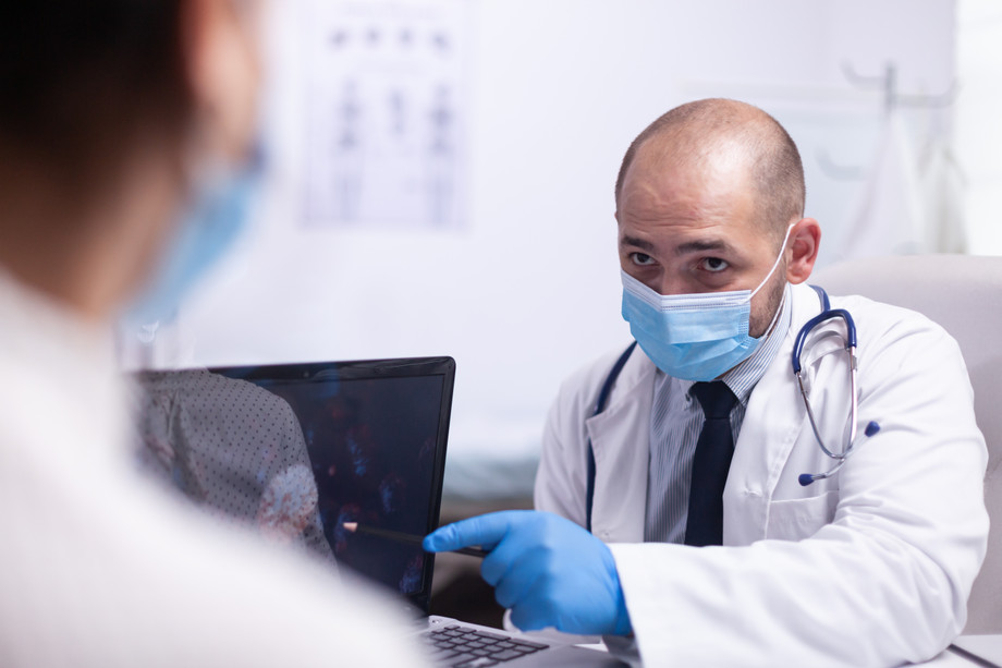 mandoctoryoungpatientwithprotectionmasktalkingaboutvirusevolutionsittingchairfrontlaptophospitalofficeclinicmedicalworkershowingsymptomspointingdesktop.jpg
