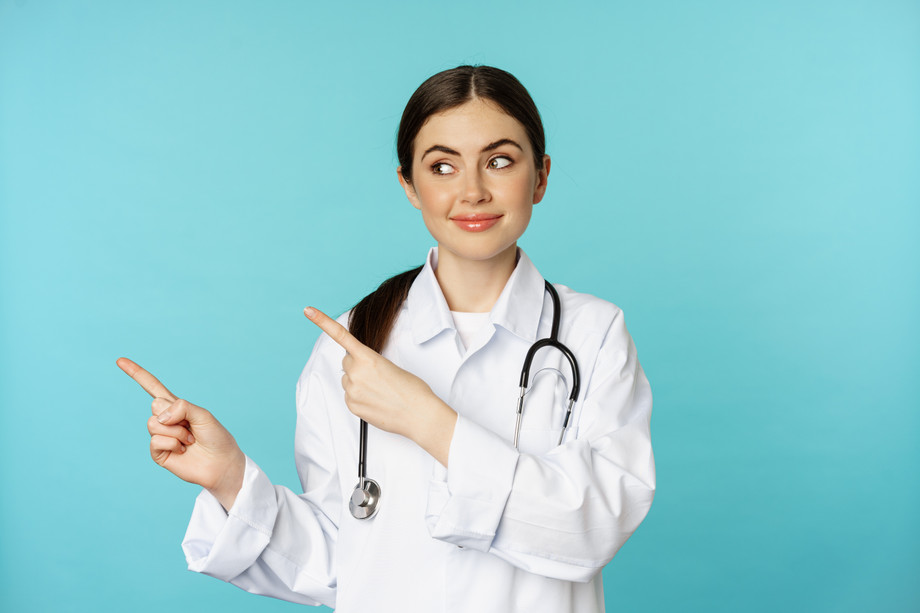 portraitsmilingmedicalworkergirldoctorwhitecoatwithstethoscopepointingfingersleft.jpg