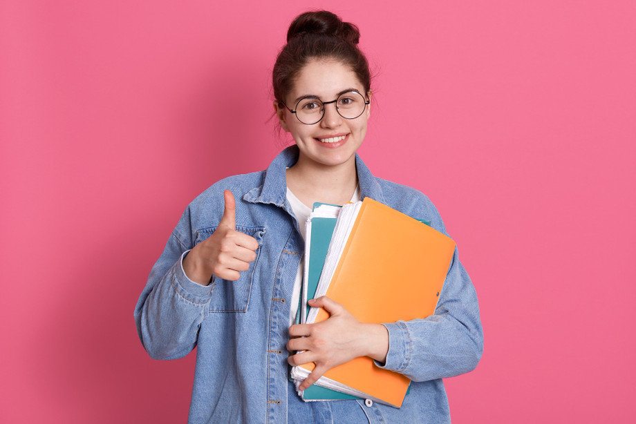 youngstudentwomanwearingdenimjacketeyeglassesholdingcolorfulfoldersshowingthumbuppink.jpg